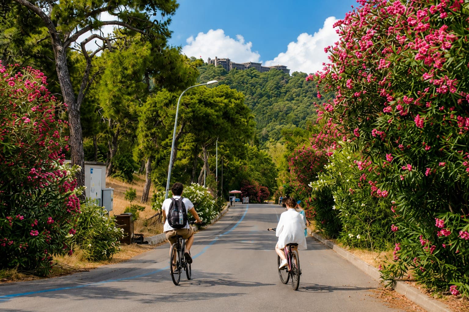Two cyclists on a flower-lined island road with a hilltop monastery beyond